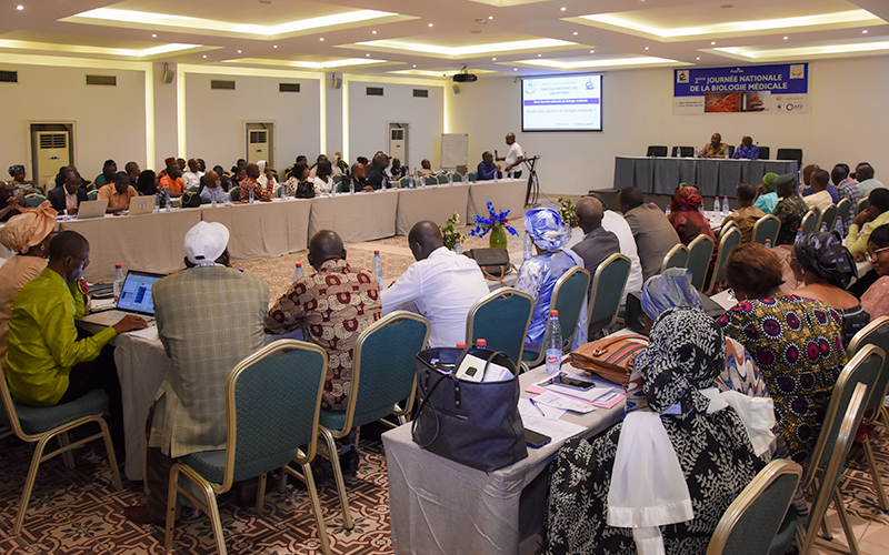 Participants at the National Medical Biology Day in a meeting room. The tables are arranged in a U shape. Opposite them is a panel and a screen.