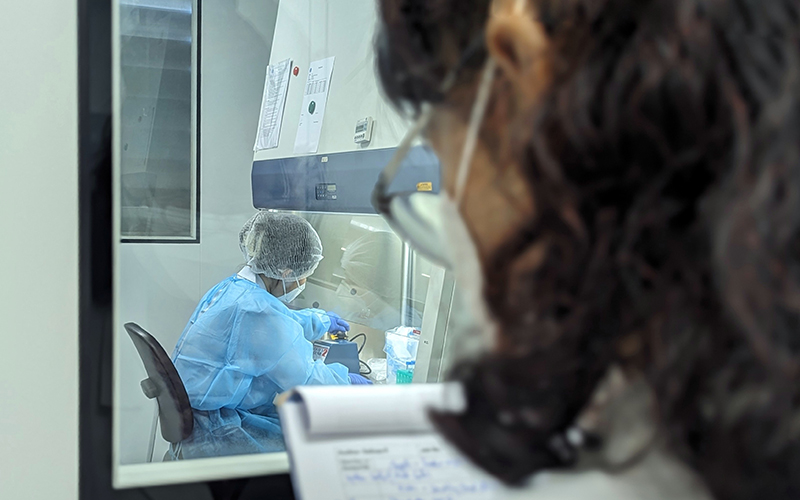 Photo taken in a laboratory. In the foreground, a person from behind, blurred, with a notebook in his hands. A window is front of this person, on the other side, another person in a lab coat is in the room, manipulating.