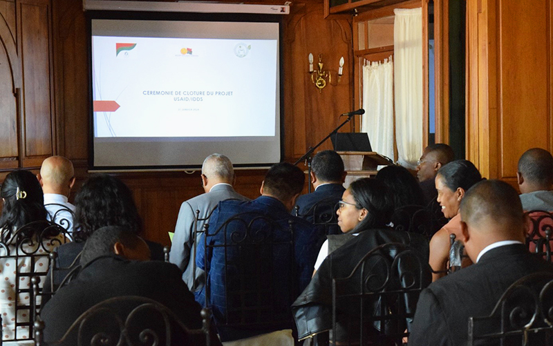 Photo of the closing ceremony of the IDDS project. Foreground: men and women with their backs to chairs, facing a lectern and a projection on the wall.