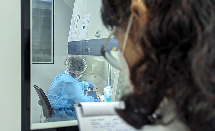 In the foreground of a laboratory, a person with a notebook in his hand. A window is front of this person, on the other side, another person in a lab coat is in the room.