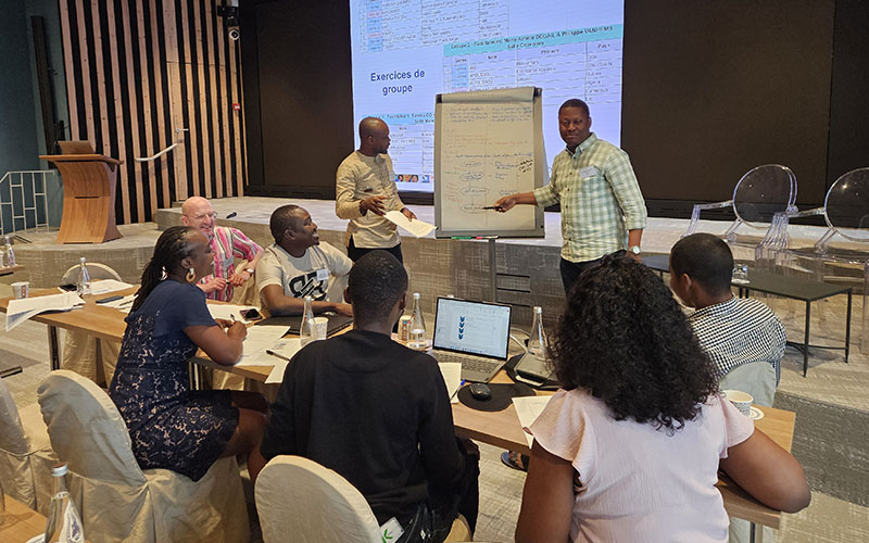 Eight people are working in a conference room. Six people are sitting in a circle, two people are standing in front of a flip chart.