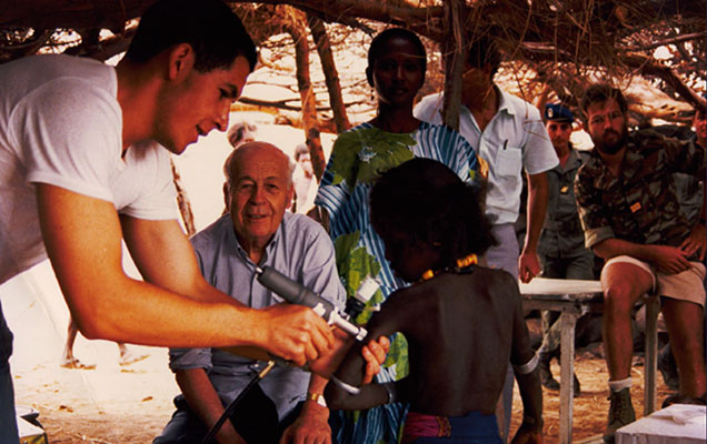 Outside, under some trees, Dr. Charles Mérieux sits and watches, in front of him in the foreground, a man vaccinating a child.