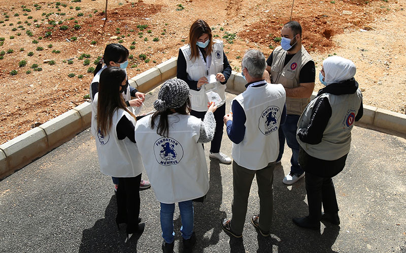 Outside, a group of eight people are standing in a circle talking. They are wearing beige vests with the blue Mérieux Foundation logo on the back.