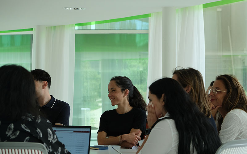 Six people are sitting around a table and looking to the left, a woman facing forward appears more clearly than the others