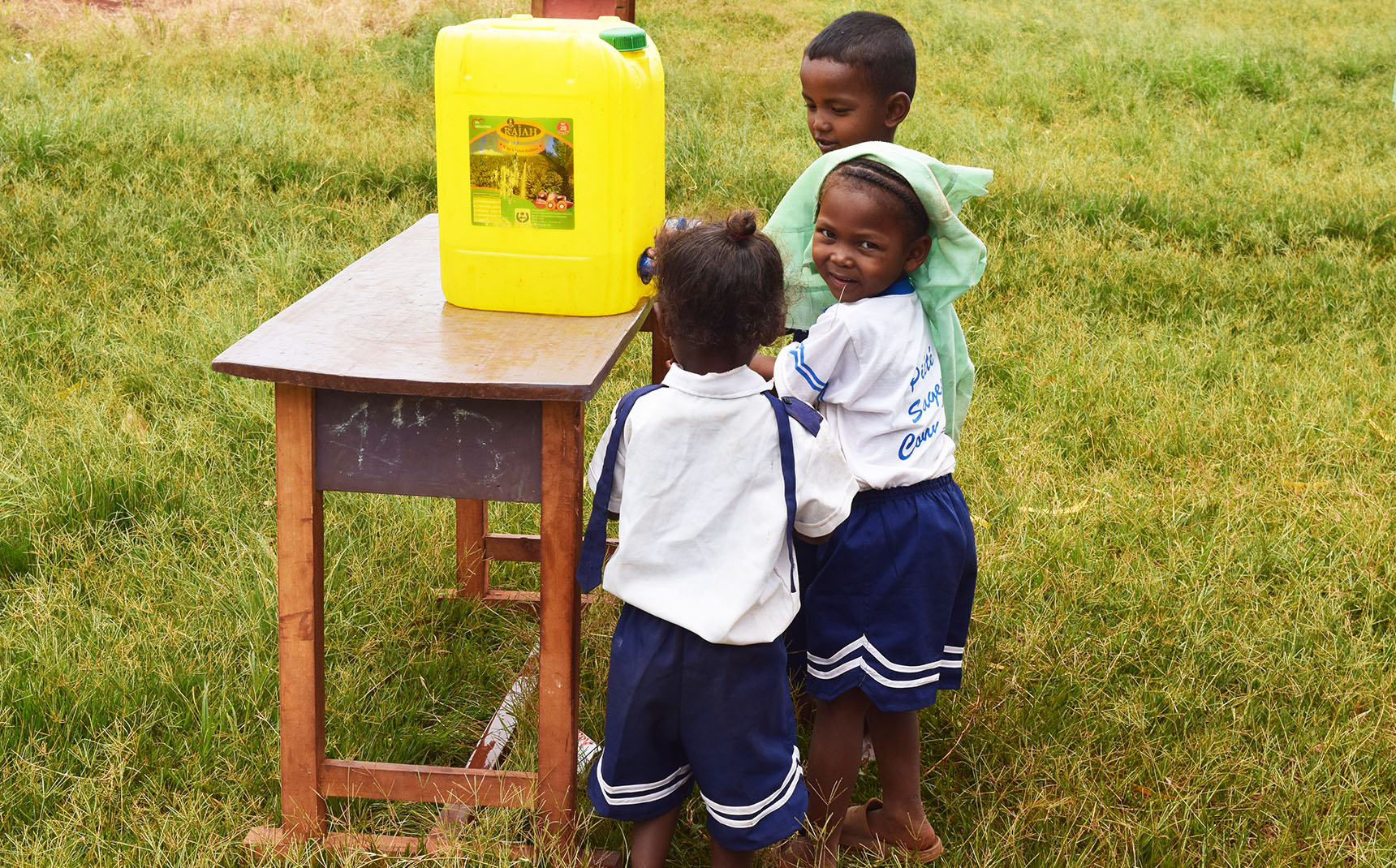 Three little Malagasy girls wash their hands from a water container.