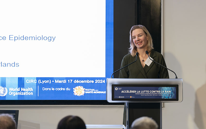 Carolien Ruesen stands at the lectern, smiling. In the foreground, the silhouettes of the audience.