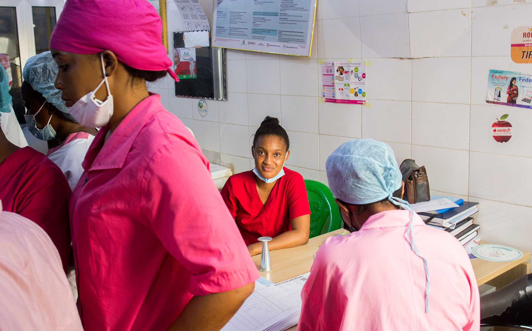 A nurse in a red coat sits facing forward and smiles, while in the foreground other nurses in pink coats are seen in profile.