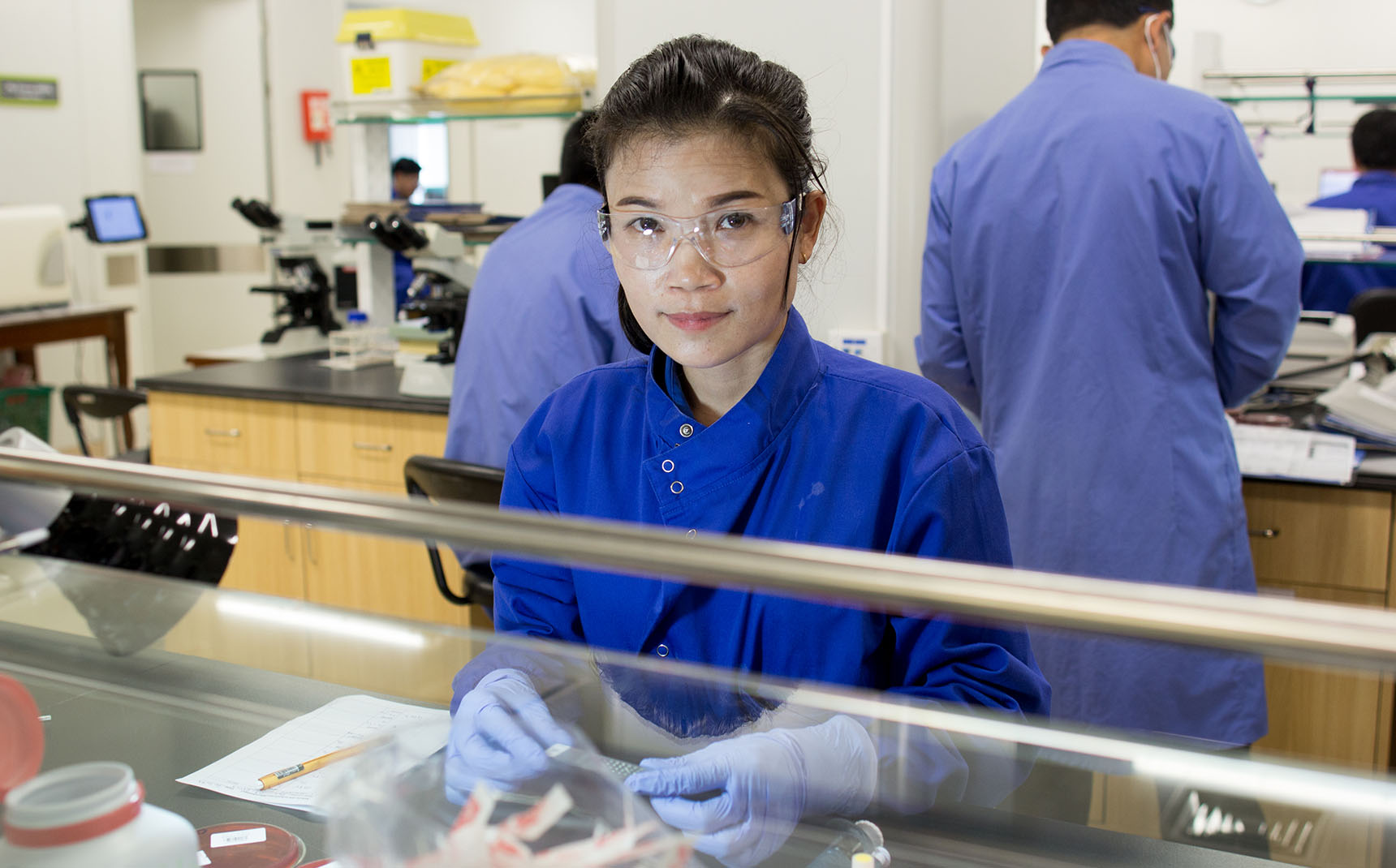 In the foreground, a lab technician in a blue coat looks at the camera, handling reagents on the lab bench.