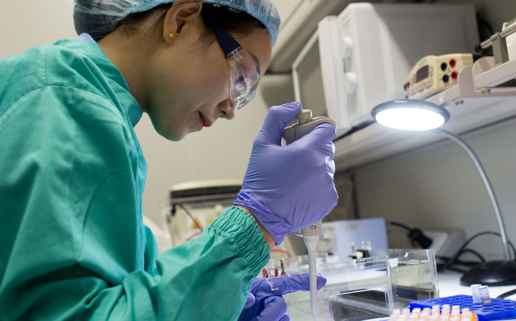 In profile, a lab technician in a green coat and green gloves manipulates a pipette.