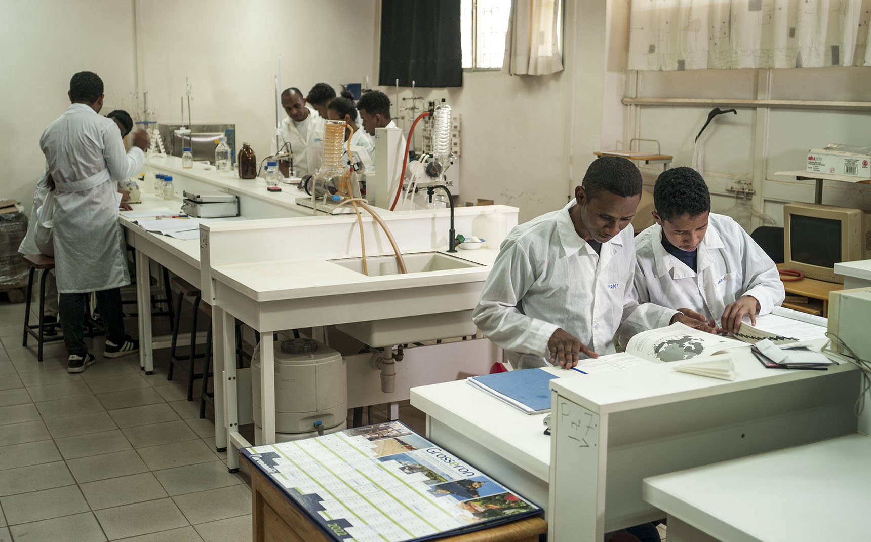 In a lab room, students are working, two students are in the foreground and a group is in the background.