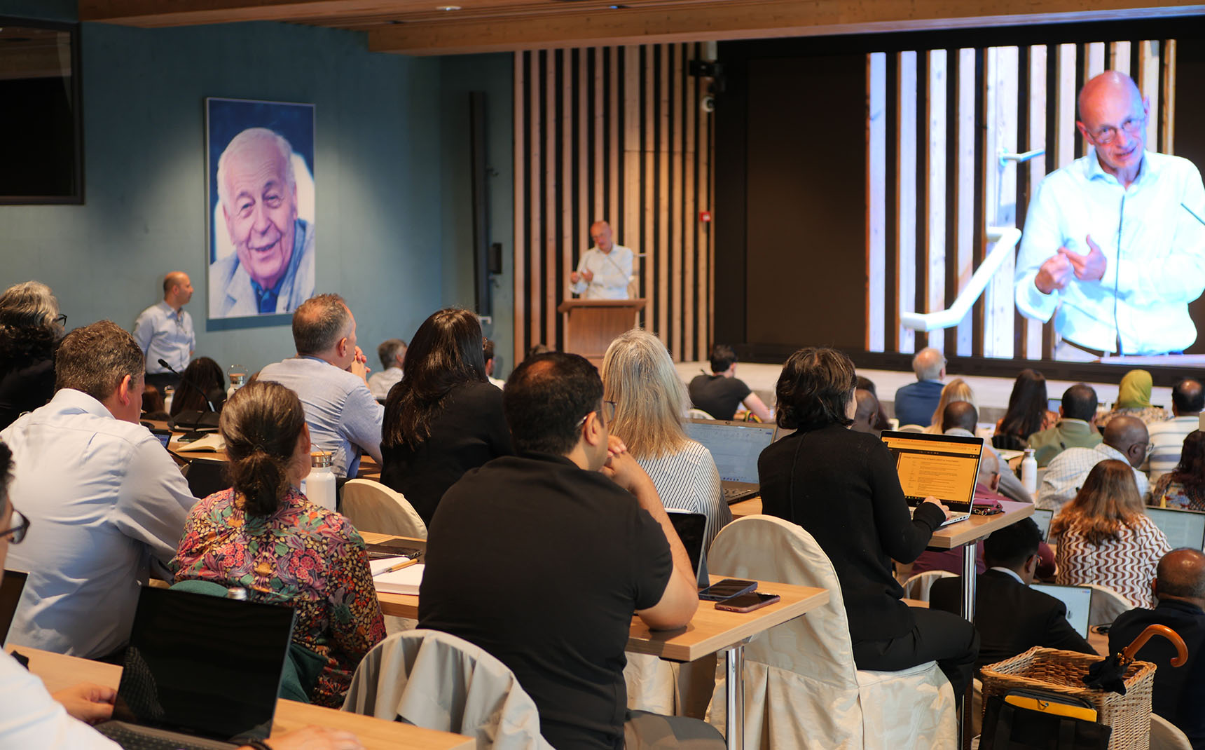 In the foreground, the audience, from behind, watches a speaker's presentation, projected onto a screen in the background.