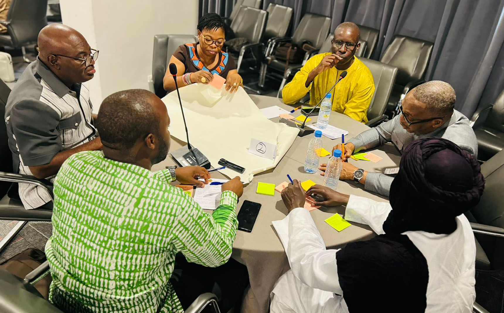 Six people sit around a round table and write on a large sheet of paper.