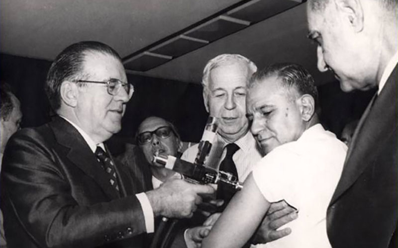 Dr. Charles Mérieux stands behind three men, one of whom is vaccinating another.