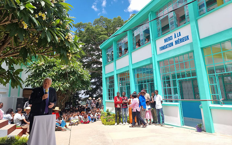 Alain Mérieux tient un micro, derrière lui sont assis des enfants et à sa droite se tient le bâtiment vert et blanc inauguré sur lequel est inscrit Merci à la Fondation Mérieux