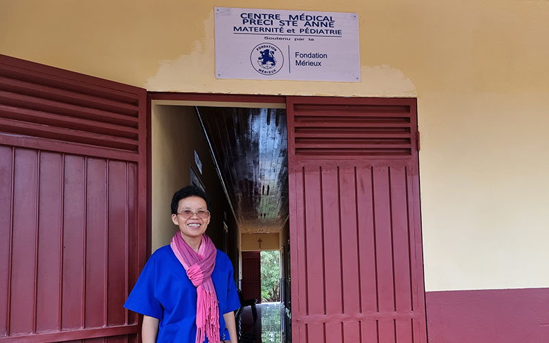 Sister Rasoafara stands on the threshold of the yellow building, above which is a plaque bearing the name of the center and the logo of the Mérieux Foundation.