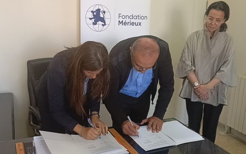 Dans un bureau, deux personnes penchées en avant pour signer des documents, en arrière plan un kakemono fait figurer le logo de la Fondation Mérieux