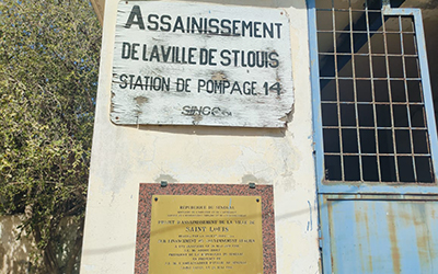 Exterior wall bearing a wooden plaque inscribed with “Assainissement de la ville de Saint-Louis” (Sanitation of the city of Saint-Louis).