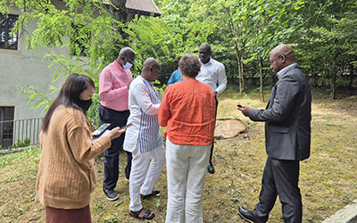 In a wooded park, a group of people are standing in a circle and working on their phones.