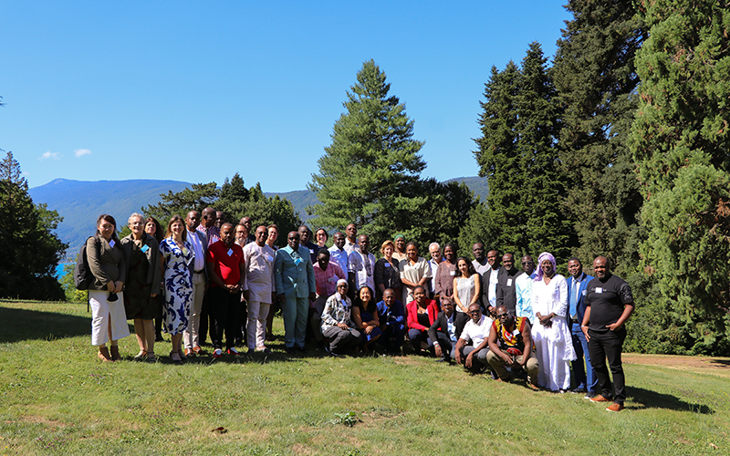 Outdoors, in a wooded park, a group of about twenty people pose standing up.