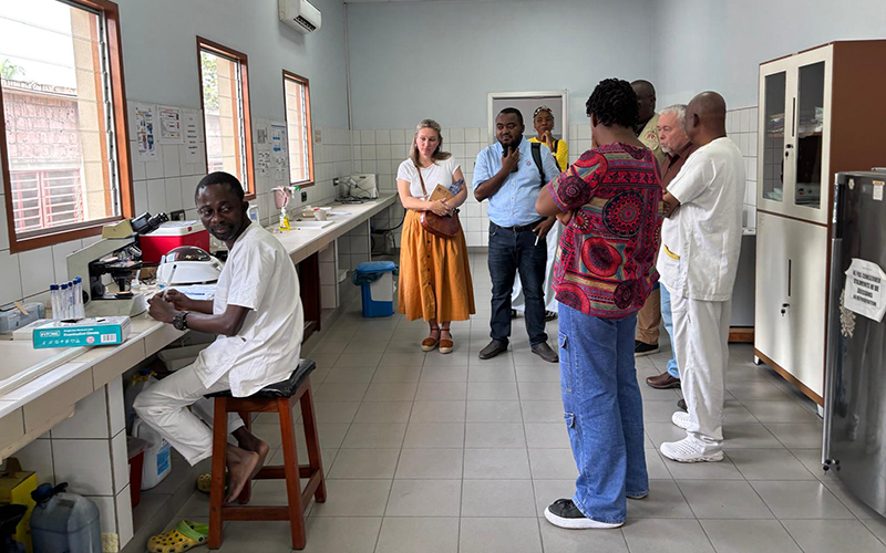 A group of people in a medical laboratory, with a technician seated at a microscope and others standing and talking. The lab is well-lit with windows, laboratory equipment on the countertops, and storage cabinets.