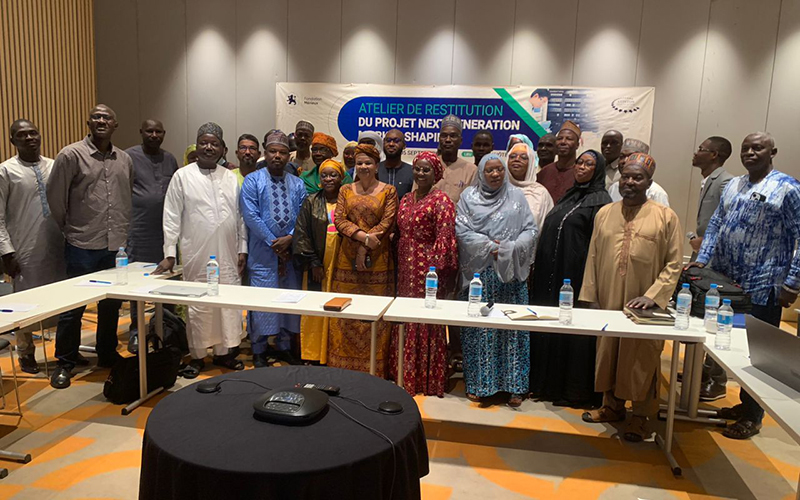In a meeting room, a group of about fifteen people stand with a blue banner in the background displaying the name of the project feedback workshop in Niger.