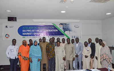 In a white meeting room, a group of about fifteen people stand in front of a blue and white banner displaying the name of the project feedback workshop in Chad.