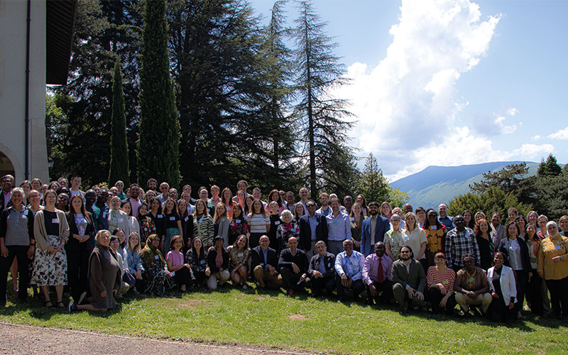 In a wooded park, a group of around 80 people pose standing on the lawn. 
