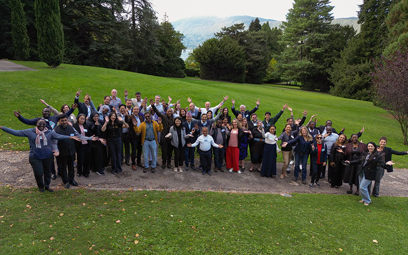 In a wooded park, a group of about thirty people pose, standing on the lawn with their arms open. 