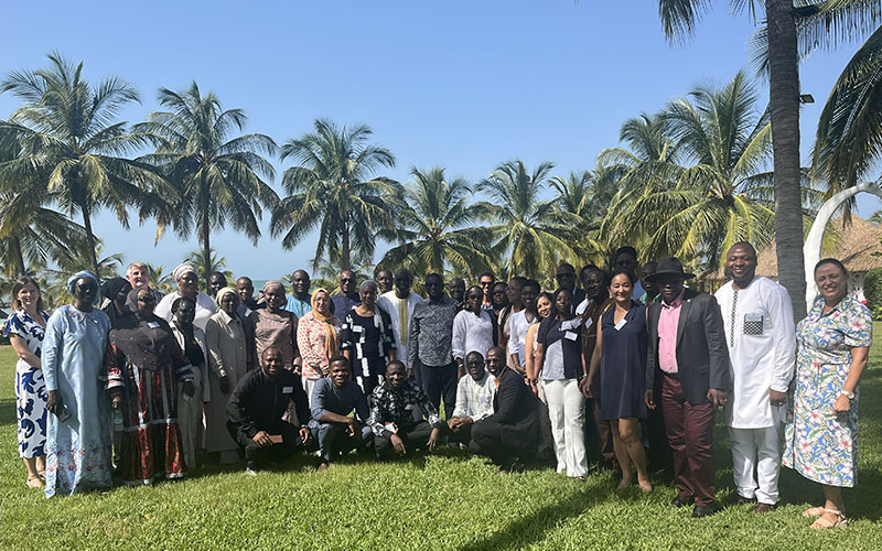Outside, on a lawn surrounded by palm trees with a big blue sky in the background and the sea visible in the distance, a group of about twenty people are standing. 