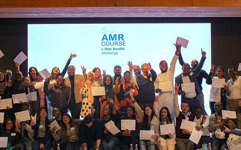On a stage in a meeting room, around thirty people pose smiling and holding up their diplomas.