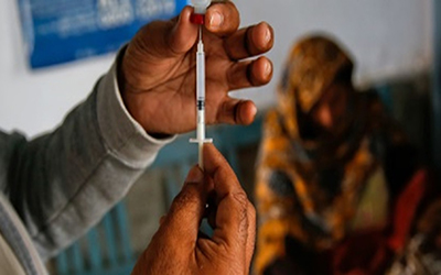 zoom sur une main gantée qui prélève à l'aide d'une seringue le vaccin contenu dans une fiole, en arrière plan flou, une femme indienne