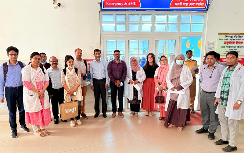 Inside, in front of the glass entrance to a hospital wing with a bright sign indicating the pediatric emergency room, a group of about fifteen individuals stands upright.