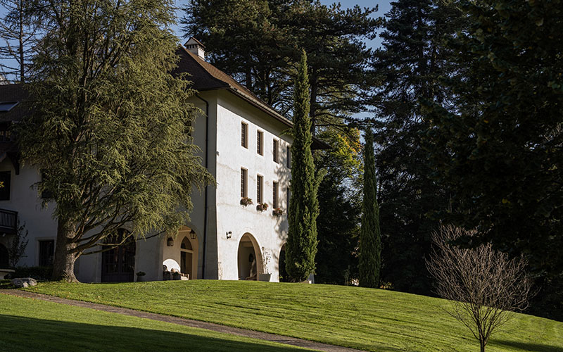 A building in typical Annecy style stands in the middle of a green lawn with many trees.