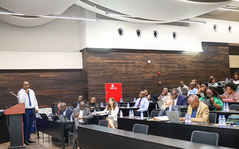 Un homme avec un micro, appuyé sur un pupitre. En face de lui, une assemblée assise écoute, regarde la présentation et prend des notes sur des carnets ou sur son ordinateur.