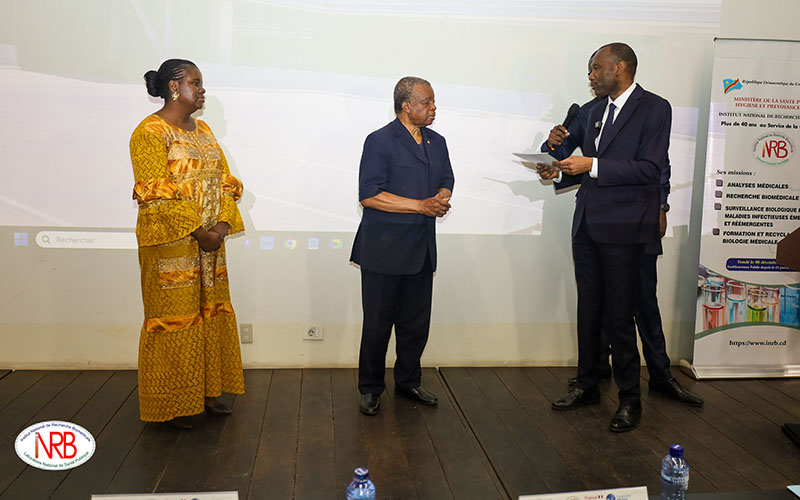 Sur une estrade en bois, trois personnes sont debout : une femme habillée en jaune regarde à droite les deux hommes. Celui de droite remet un document papier à celui du milieu