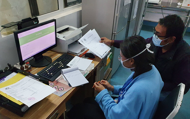 Deux personnes se trouvent devant un écran d'ordinateur qui est allumé et projette le logiciel. L'écran se trouve sur un bureau, avec une imprimante et des carnets.