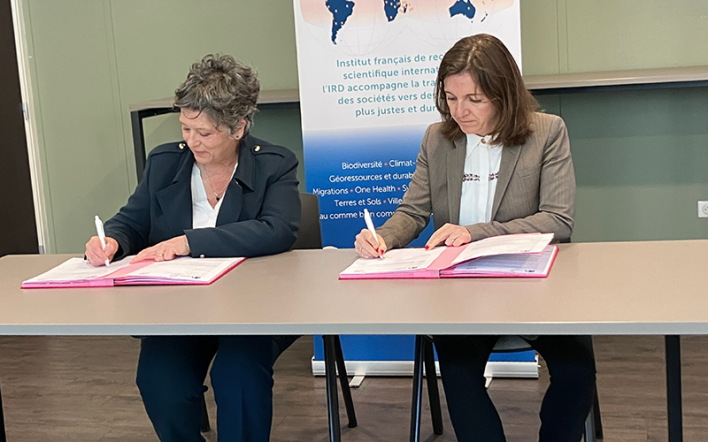 Two women are sitting down, with a table in front of them. They have some documents in front of them, and they are signing them.
